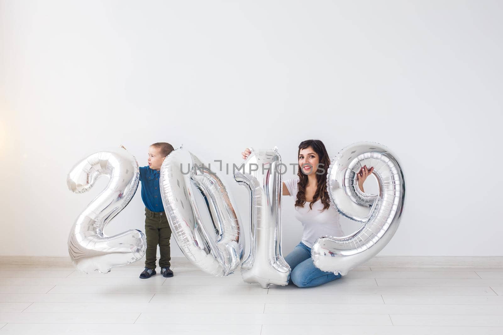 New year, celebration and holidays concept - mother and son sitting near sign 2019 made of silver balloons for new year in white room background.