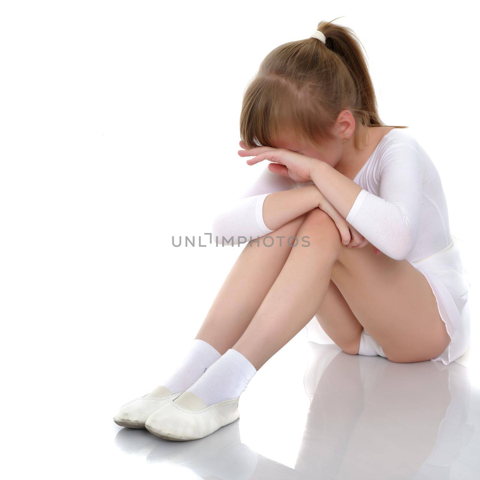 A sweet little gymnast girl is upset, she sits on the floor and cries. The concept of sport, children's emotions. Isolated on white background.