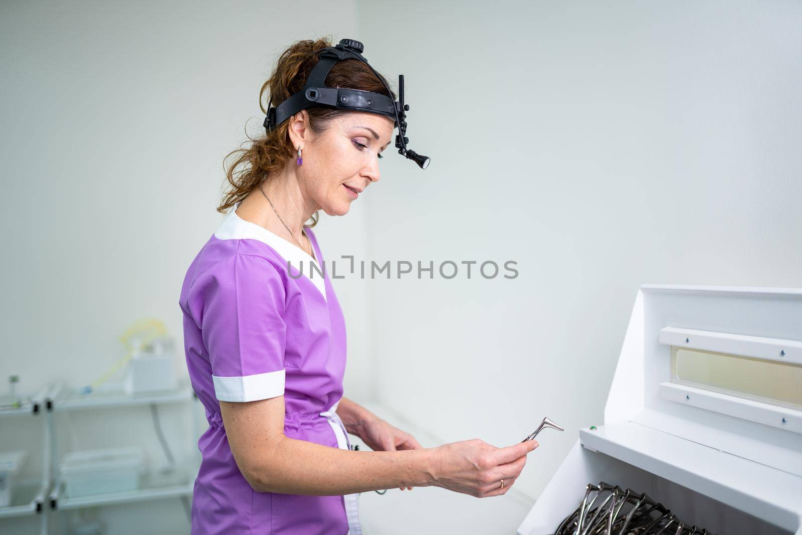 Medical worker of ENT clinic middle aged Caucasian female in purple medical uniform posing looking at the camera in the examination room. Professional medical specialist otolaryngologist in hospital by Tomashevska