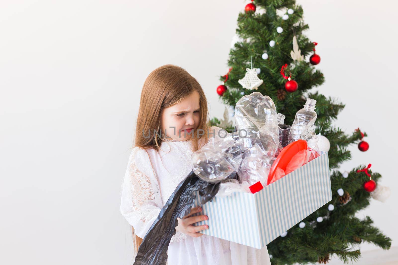 Shocked little child girl looks with opened eyes and worried expression, holding box with various plastic wastes over christmas tree background