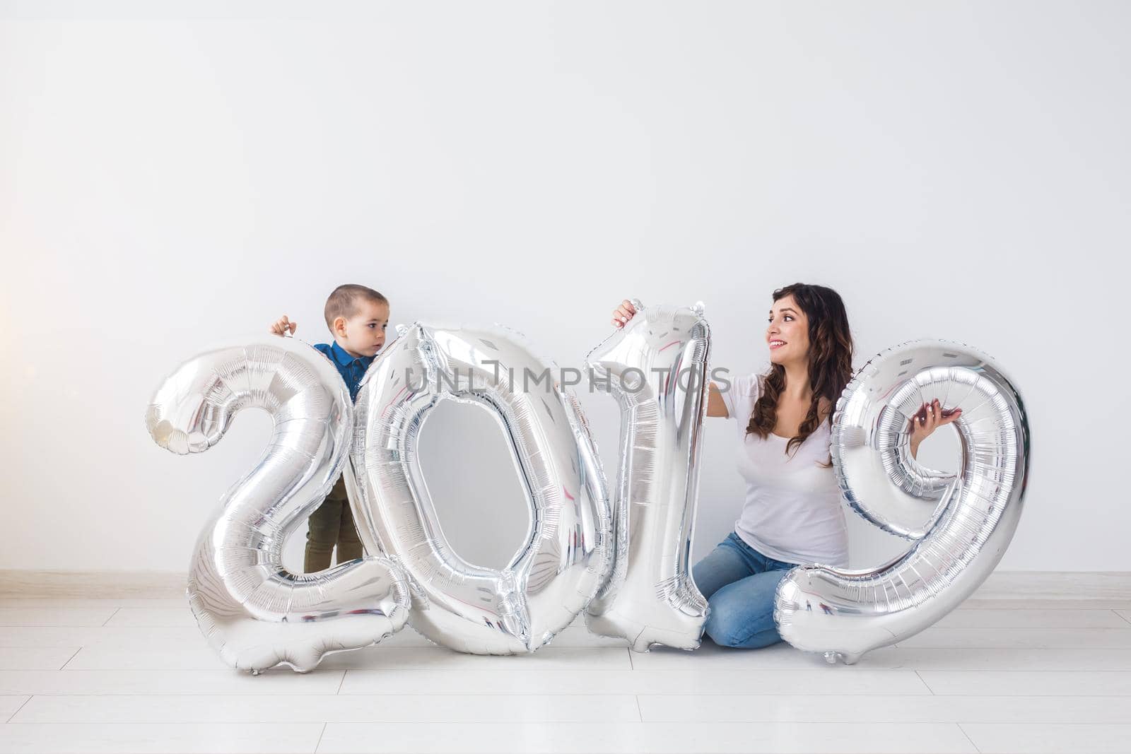 New year, celebration and holidays concept - mother and son sitting near sign 2019 made of silver balloons for new year in white room background by Satura86