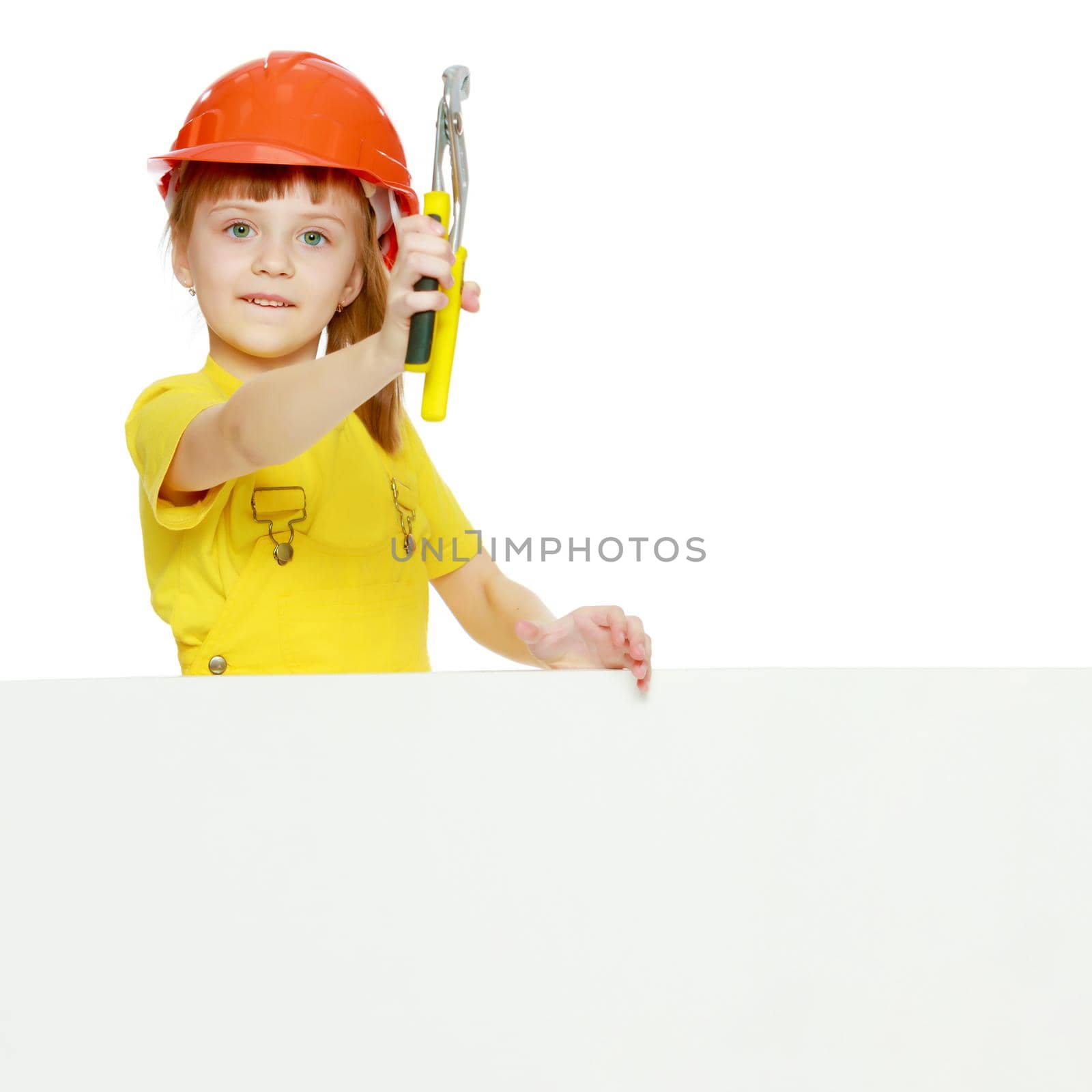 A nice little girl in a construction helmet and a yellow T-shirt peeped out from behind a white advertising banner.Girl in a construction helmet peeks out from behind a billboard.