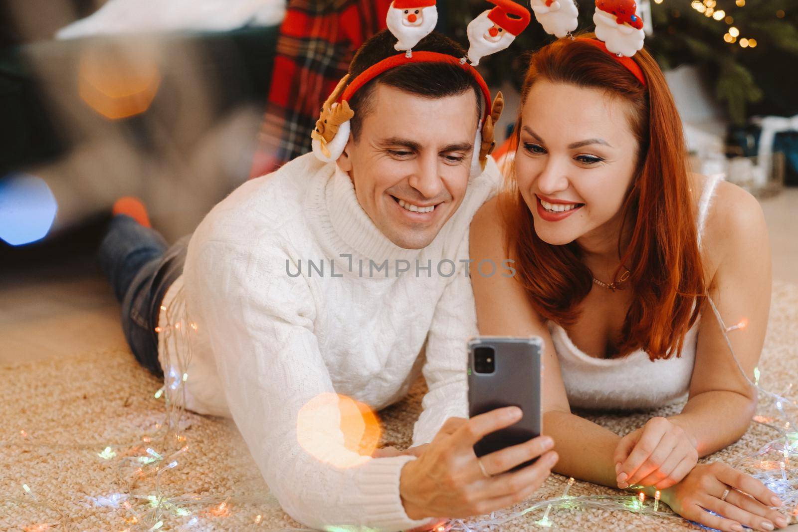a happy married couple is lying on the floor at home near the Christmas tree and taking pictures of themselves by Lobachad