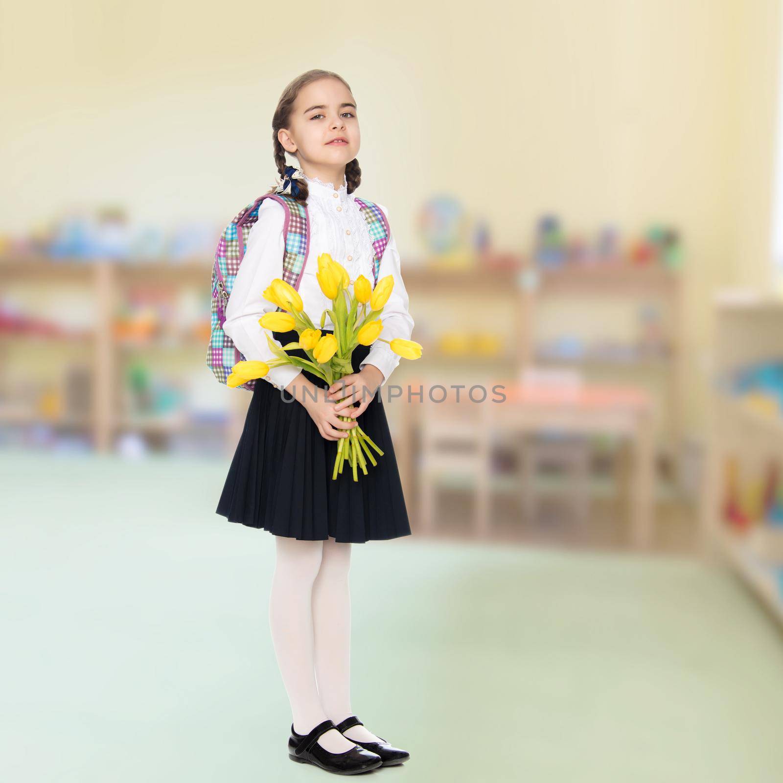 Beautiful little blond schoolgirl, with long neatly braided pigtails. In a white blouse and a long dark skirt.She is holding a bouquet of yellow tulips.In the Montessori Room the children's garden.