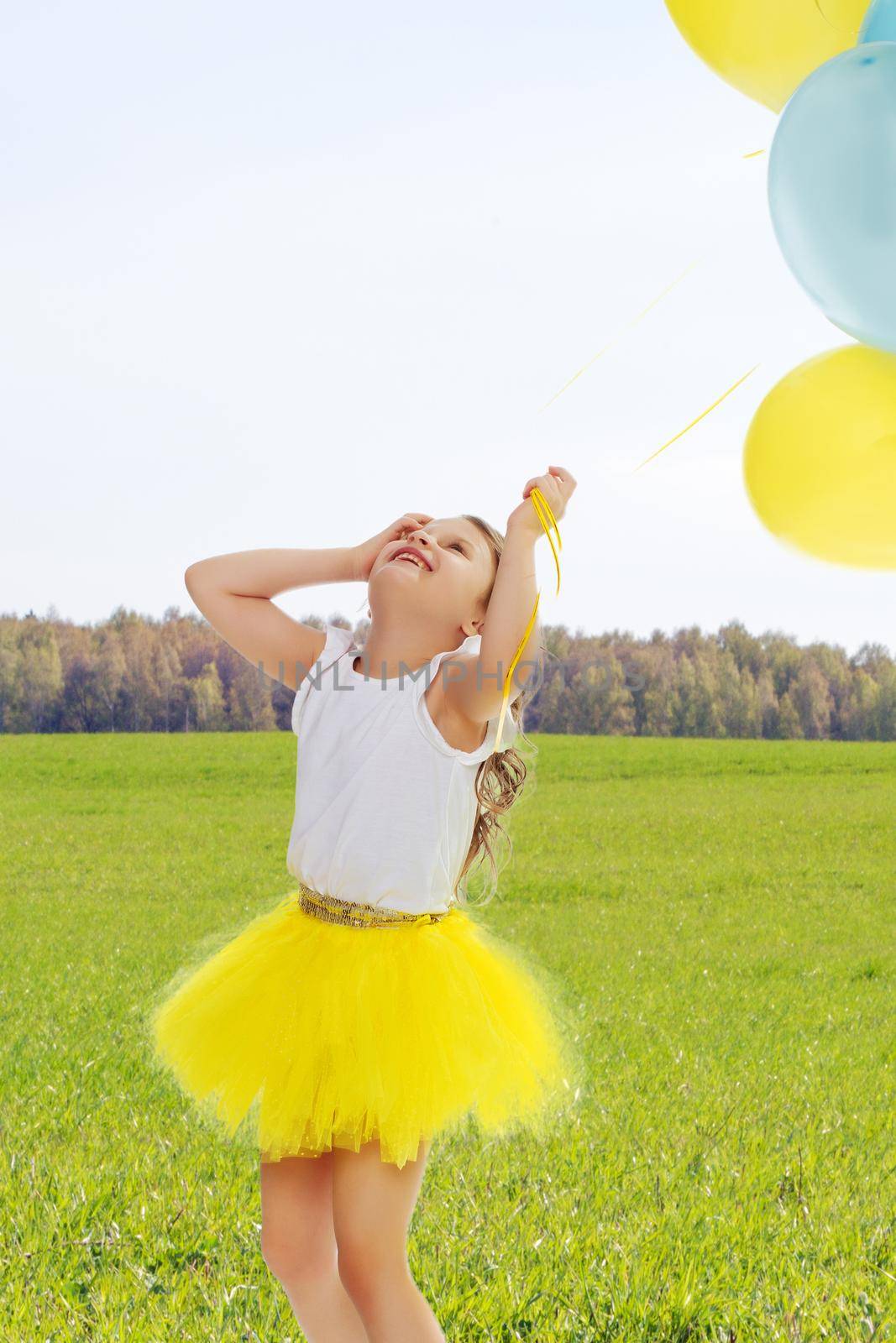Pretty little blonde girl in a short bright yellow skirt and white blouse.Girl holds in hands balloons , She looks at them from the bottom up.On the background of green grass and blue sky with clouds.