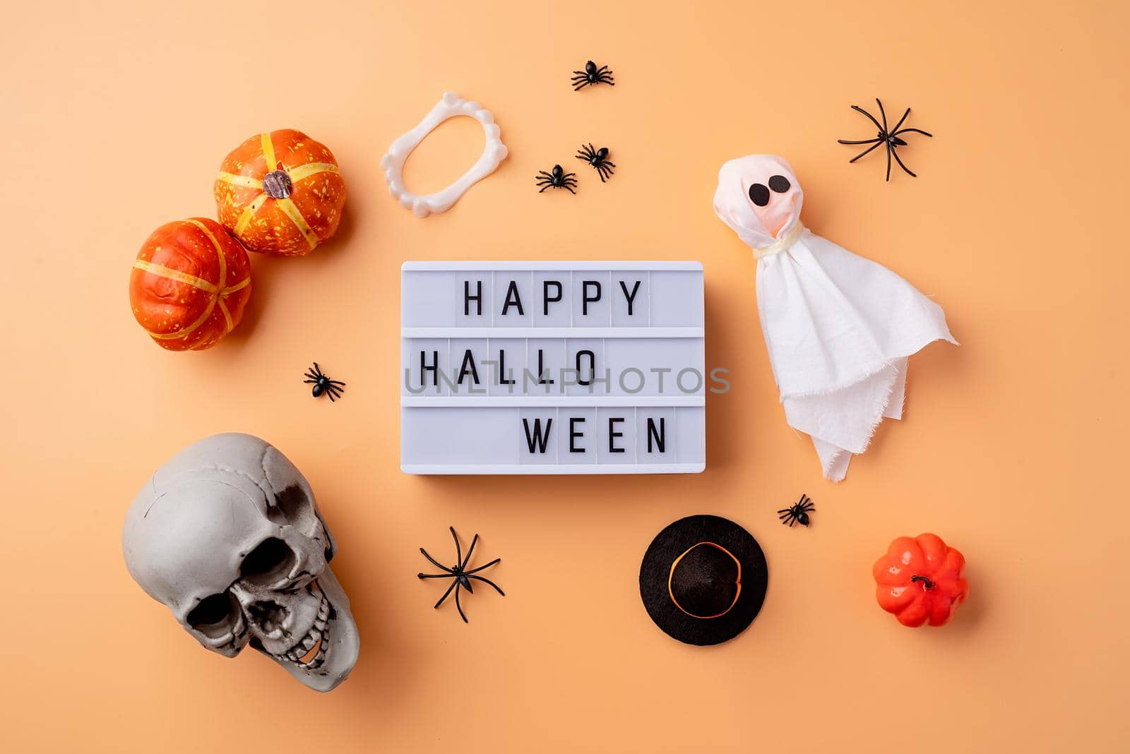 Halloween concept. Halloween party decorations with lightbox with words Happy Halloween, scull, pumpkins, ghost and spiders top view flat lay on orange background