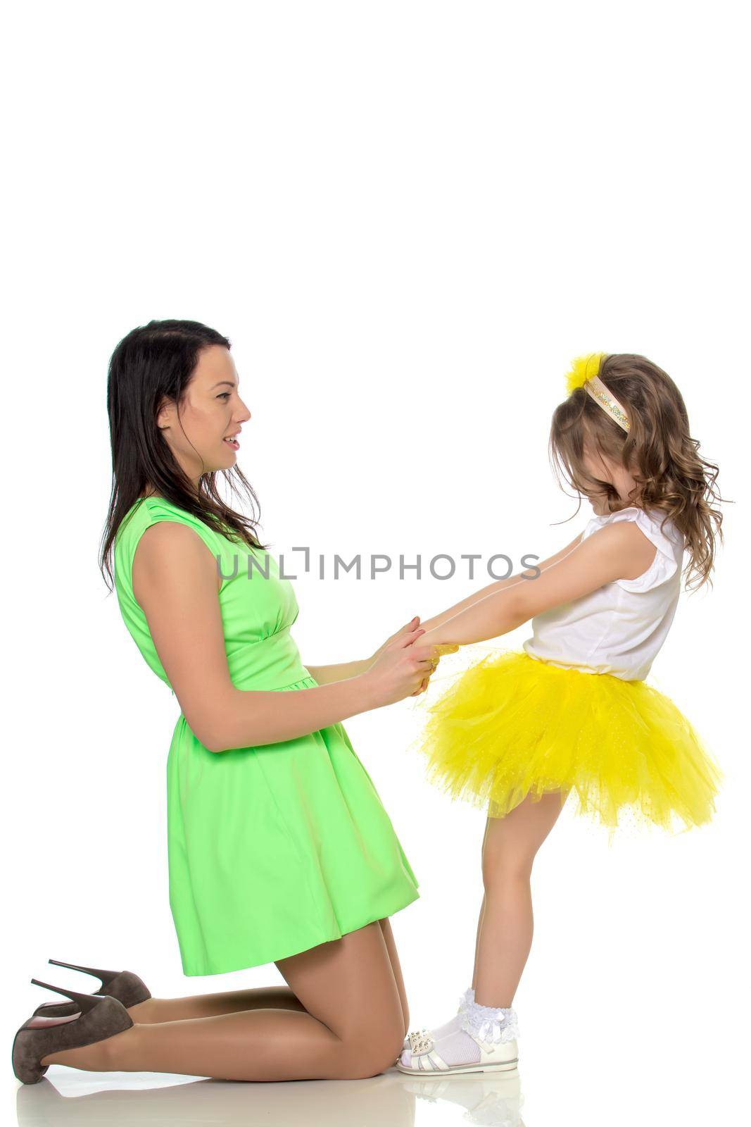Beautiful young mother in a short green dress and her little beloved daughter in a yellow skirt.They have fun holding hands.Isolated on white background.