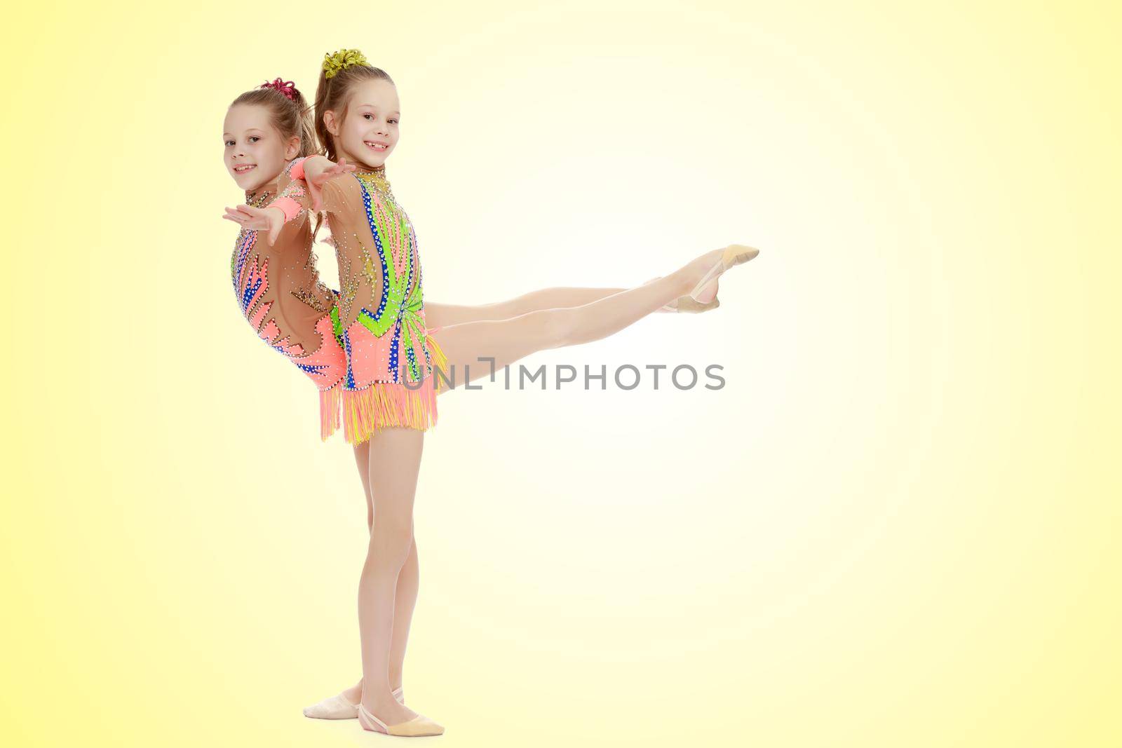 Two adorable little twin girls, gymnastics in the sports school. Girls beautiful gymnastic leotards. They do the splits.On a yellow gradient background.