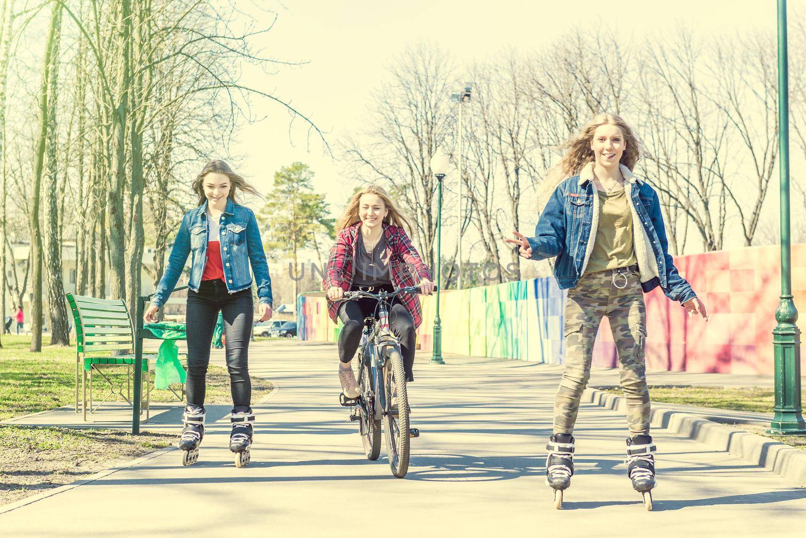 Group of three teen girls in clourful trendy clothes having fun while roller skating and riding a bicycle at park