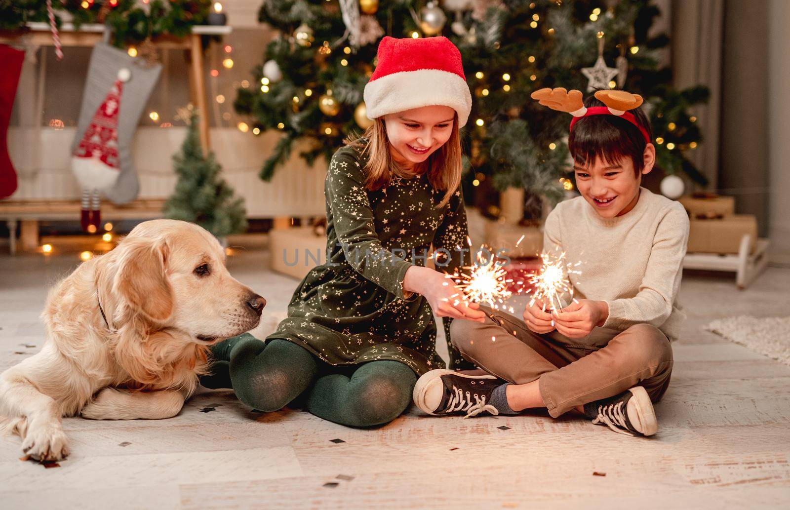 Little girl in santa hat and boy wearing reindeer horns rim holding sparklers while sitting beside golden retriever dog at home