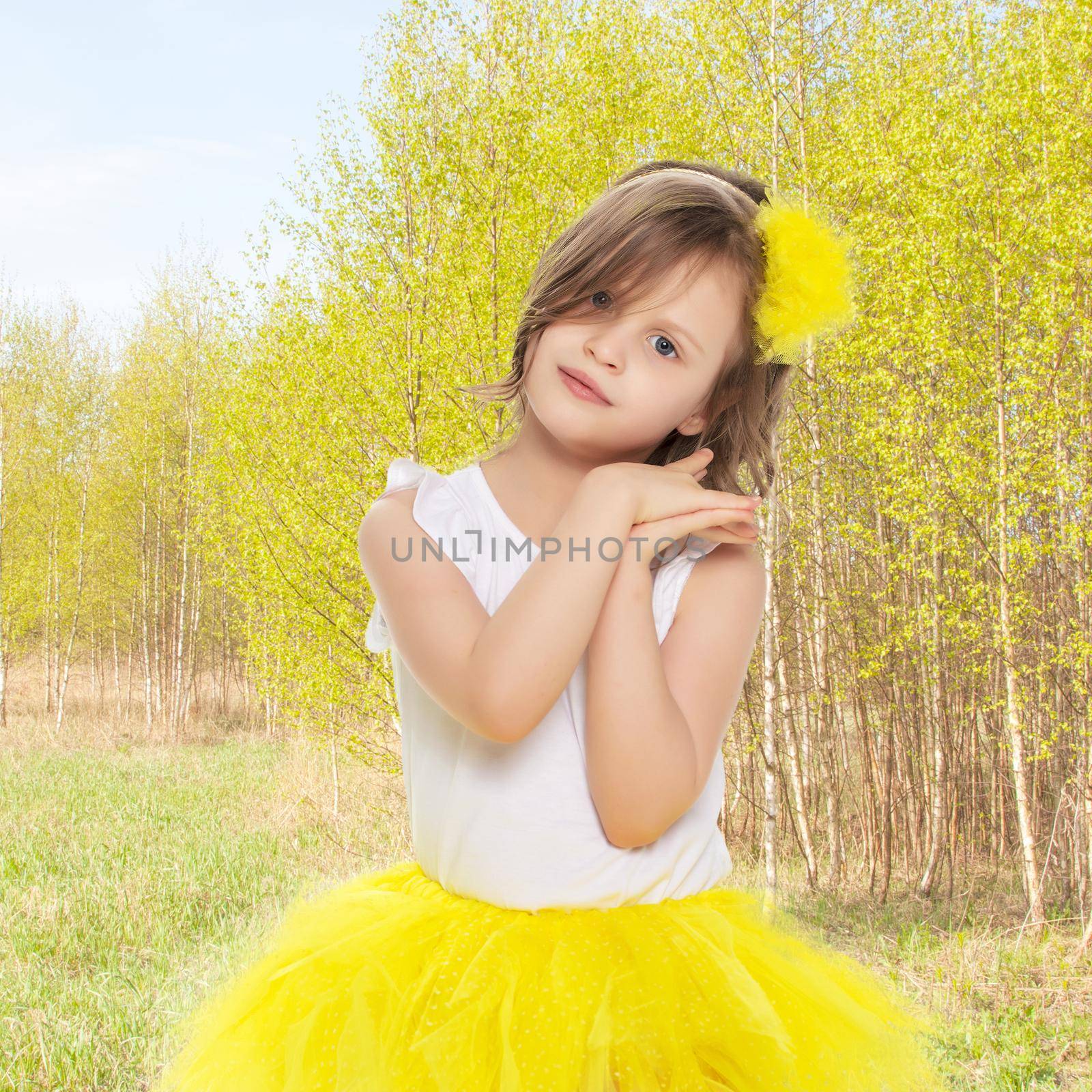 Pretty little blonde girl in a short bright yellow skirt and white blouse.She kneels and gently looking at the camera with folded hands near the face.Against the background of the summer landscape.