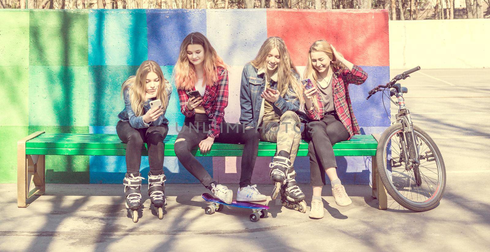 Trendy young girls laughing while using mobile phones in a skateboard park. Four pretty girl friends sitting on the bench with skateboards, rollers and bicycle