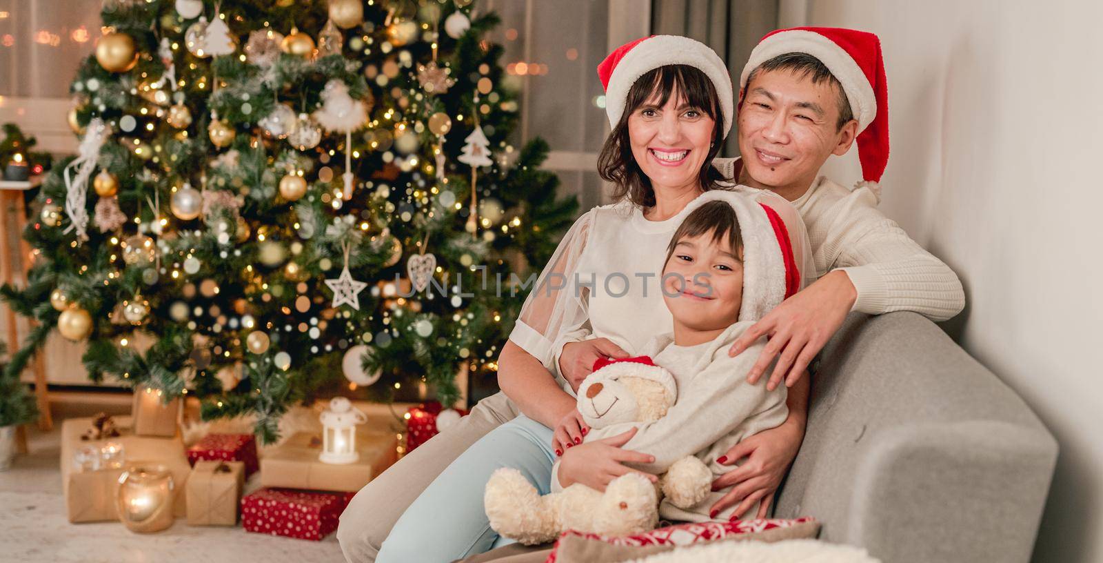 Happy family in santa hats sitting on floor under decorated christmas tree at home