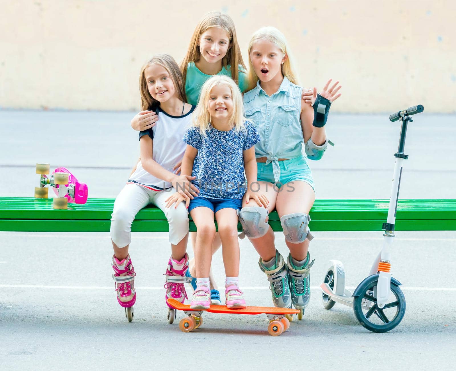 Pretty smiling little girls sitting hugging on the green bench in skatepark with scooter,skates and rollerskates put on