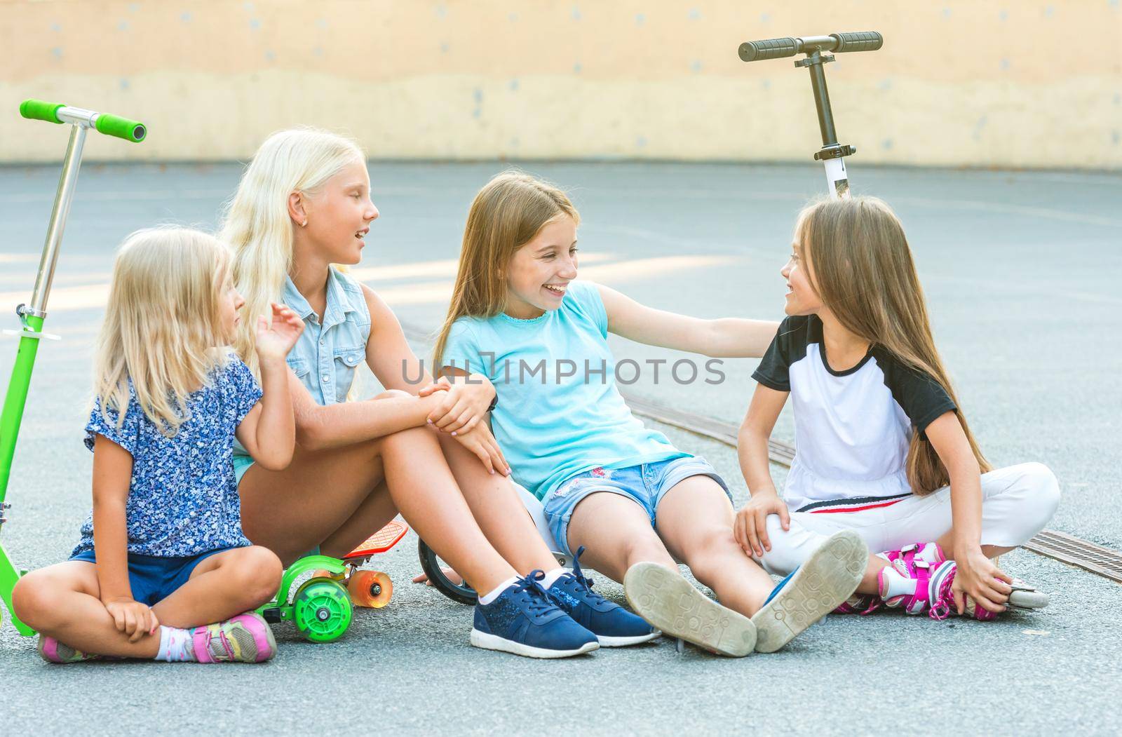 Pretty Little girls talk sitting on the ground with scooters ,skate and rollerskates put on
