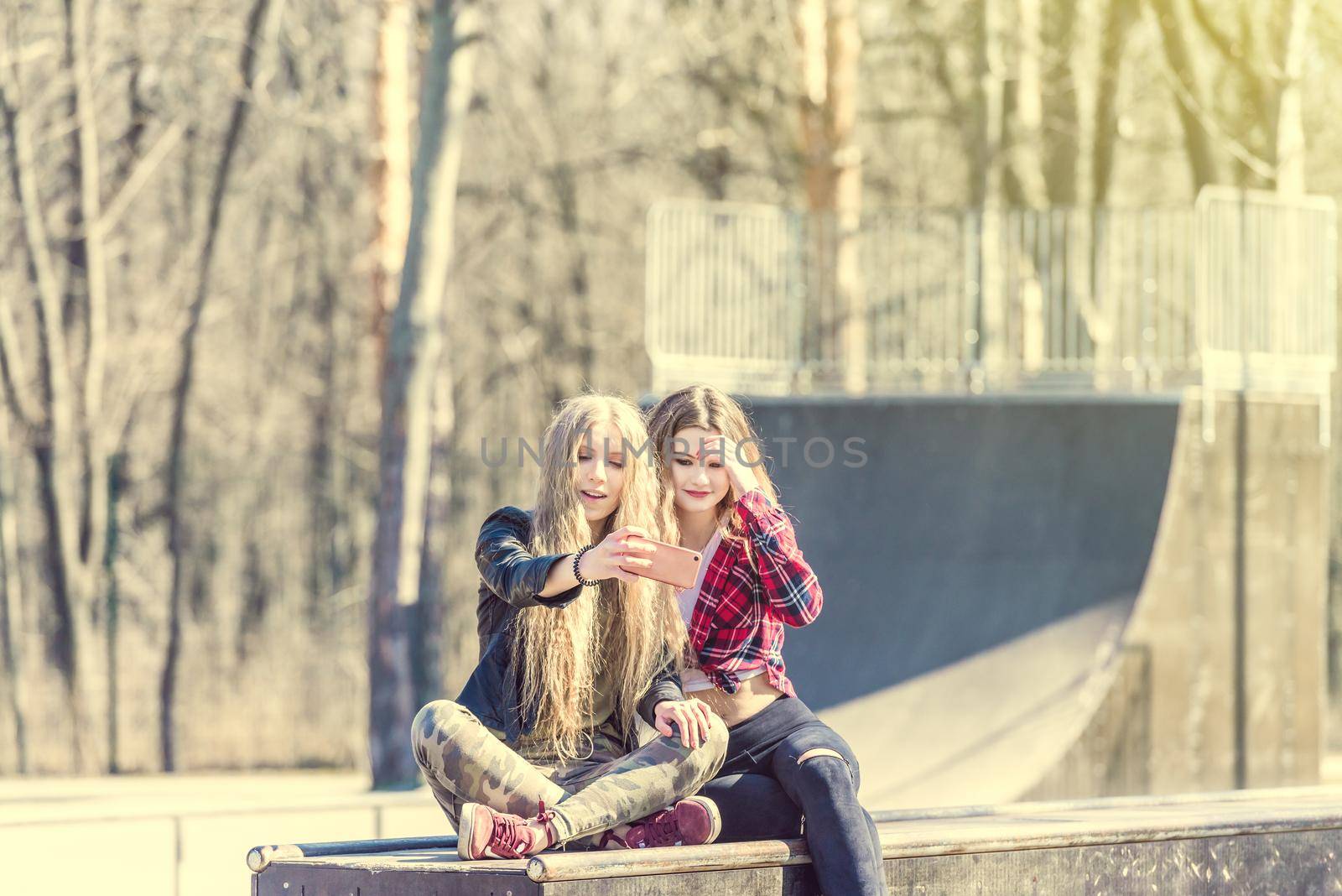 Two beautiful girls in casual clothes doing selfie sitting on ramp at skate ground at sunny day. Best female friends hanging out at skate park