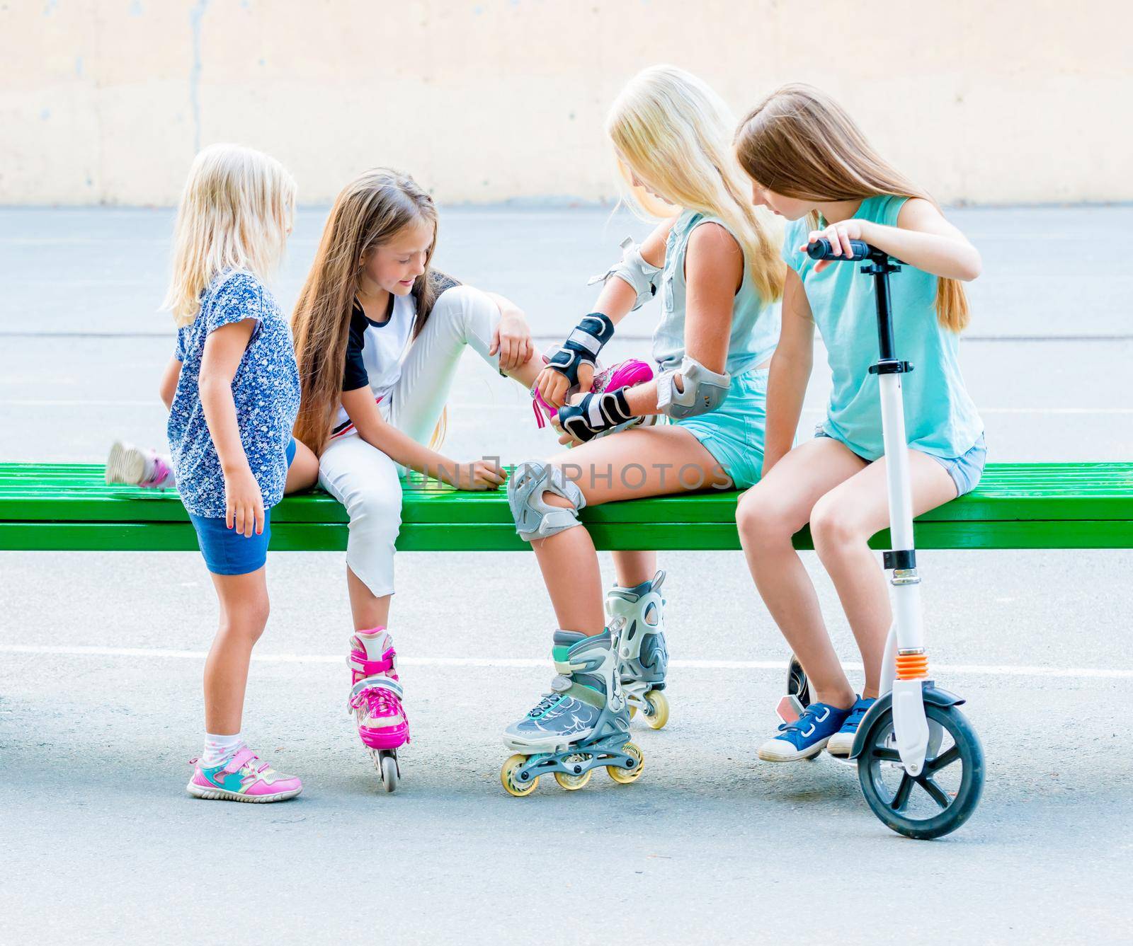 Little girls sitting on the green bench putting on rollerskates