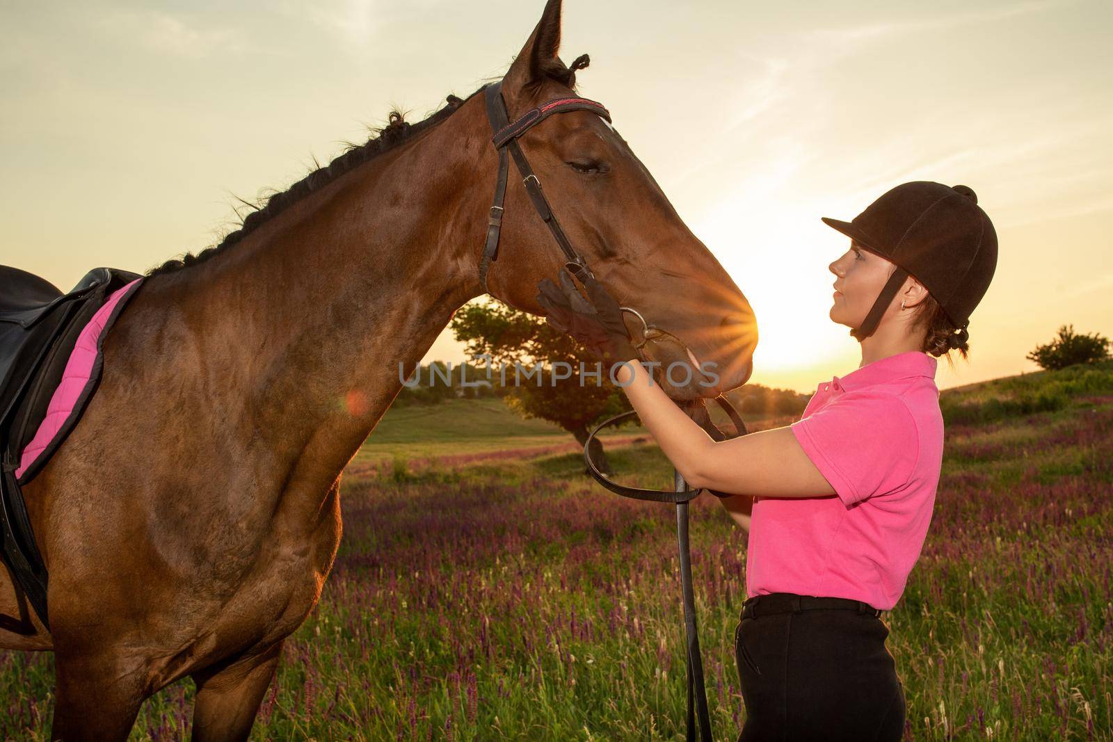 Beautiful smiling girl jockey stand next to her brown horse wearing special uniform on a sky and green field background on a sunset. Equitation sport competition and activity.