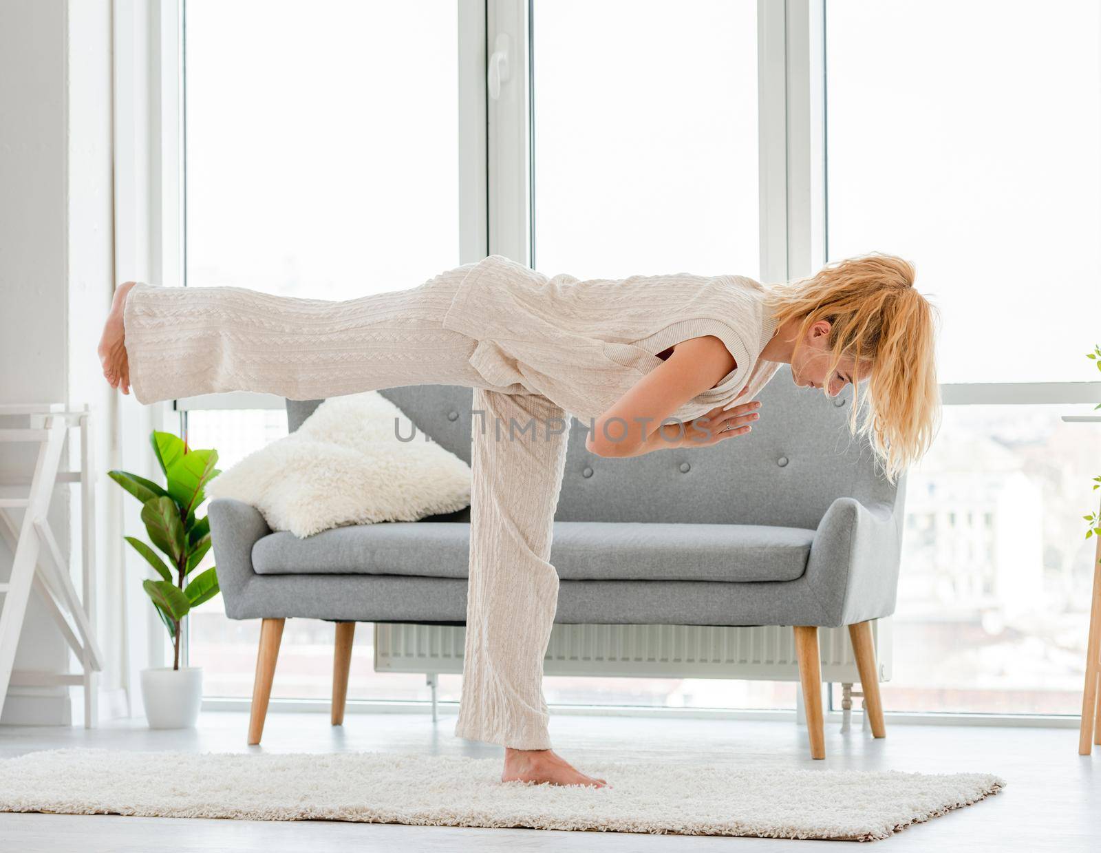 Blond girl keeps balance standing on one leg during morning yoga workout at home