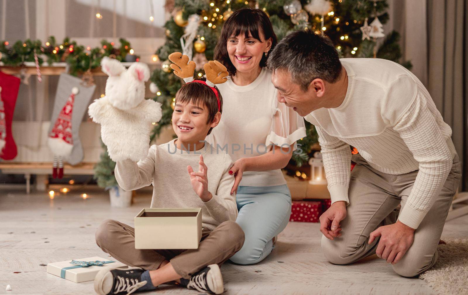 Happy little boy holding plush toy from christmas present sitting with parents under new year tree at home