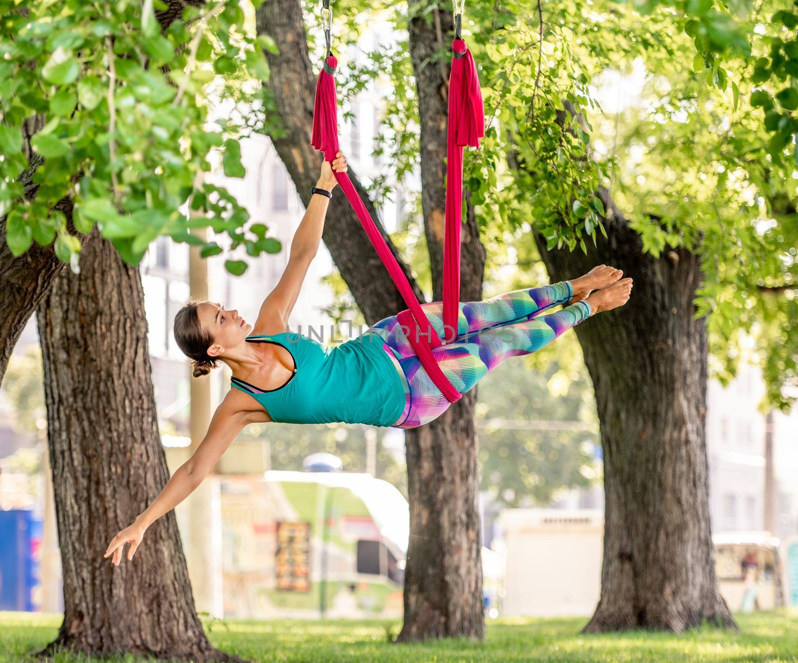 Girl practicing fly yoga in hammock and stretching her body at nature with balance in the air. Young woman doing aero fitness gymnastic exercising outdoors in summer