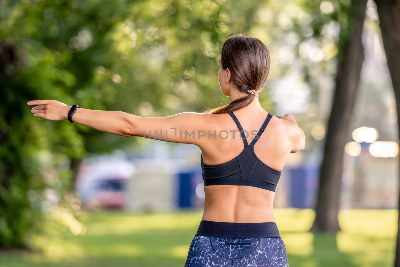 Sport girl doing yoga workout at nature in the morning. Young woman exercising and stretching arms outdoors portrait from back