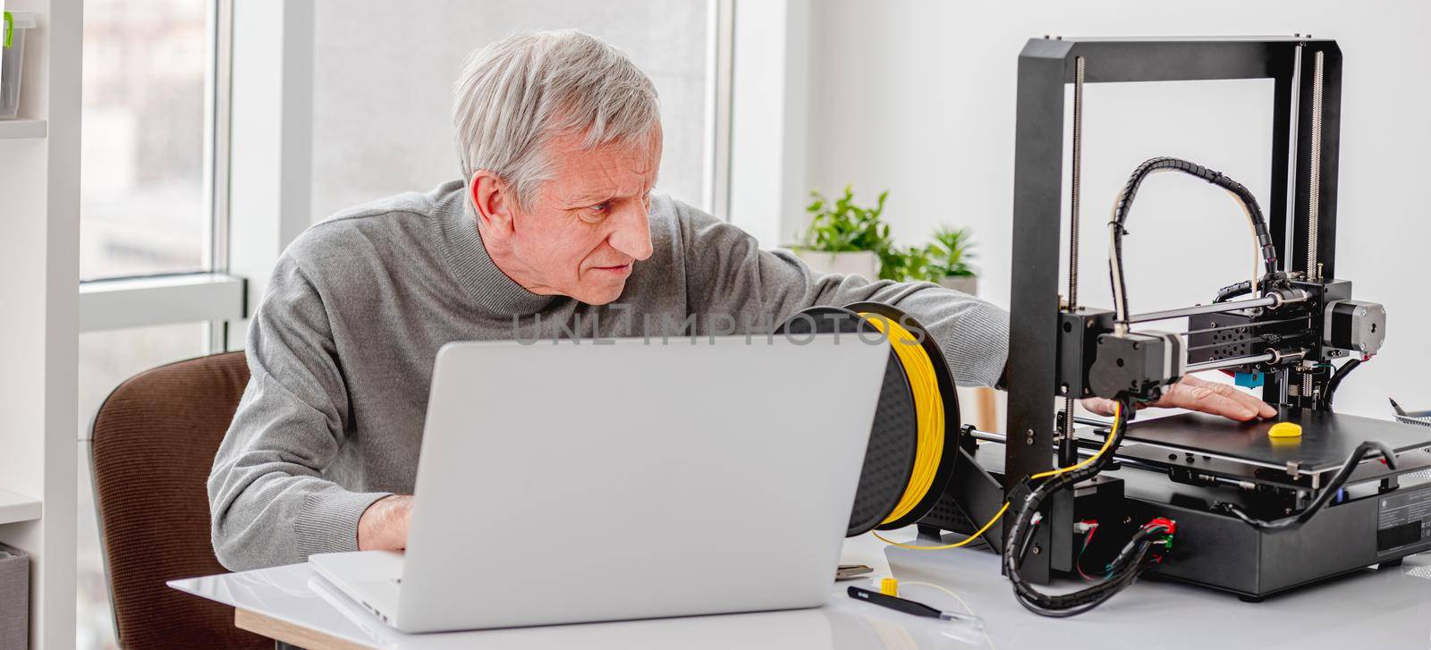 Adult man working on design project with 3D printer and laptop sitting by the table at office