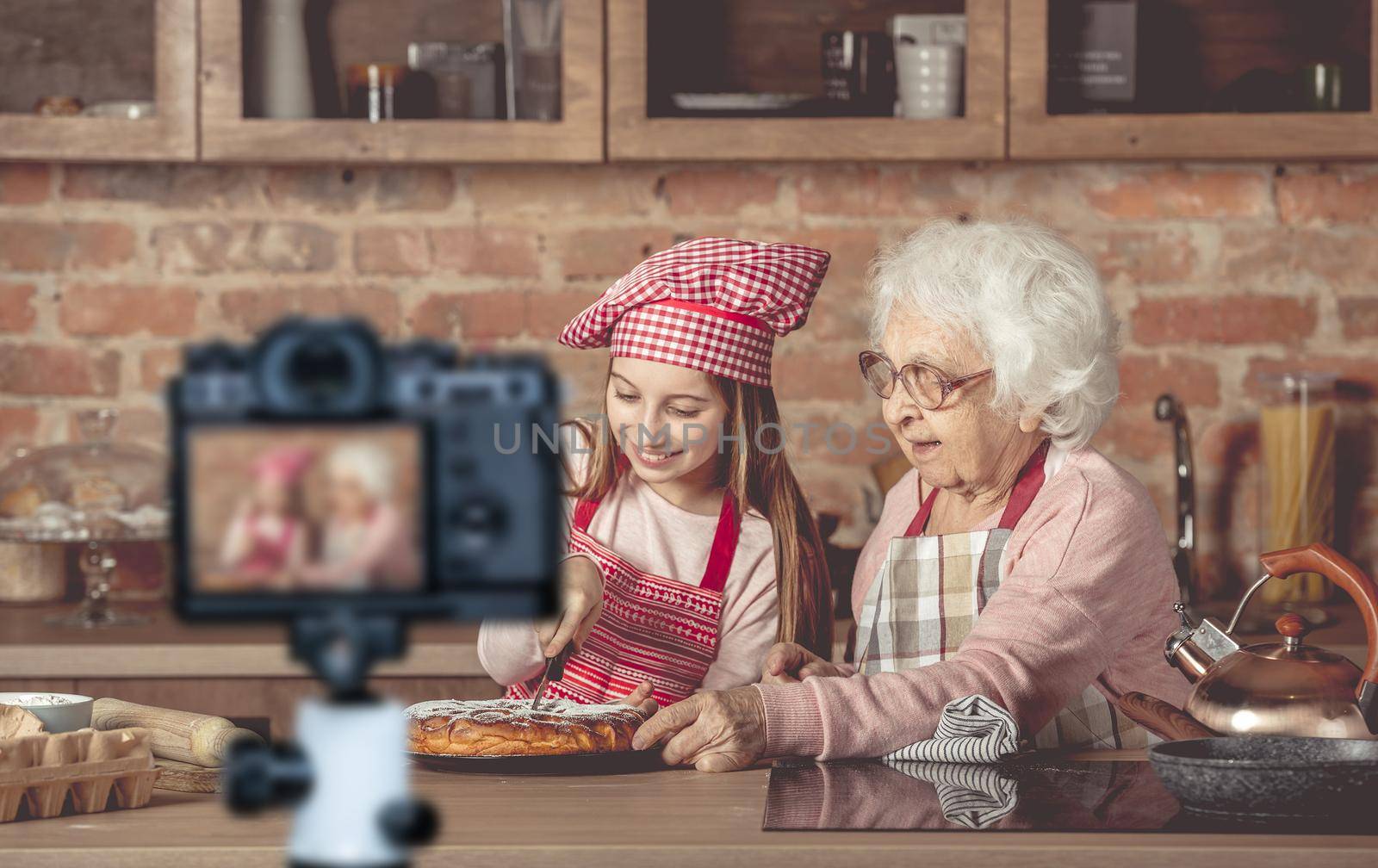 Granddaughter with her grandmother sitting at the kitchen and cut the homemade pie. Blogger profession. Shooting with a camera for a blog