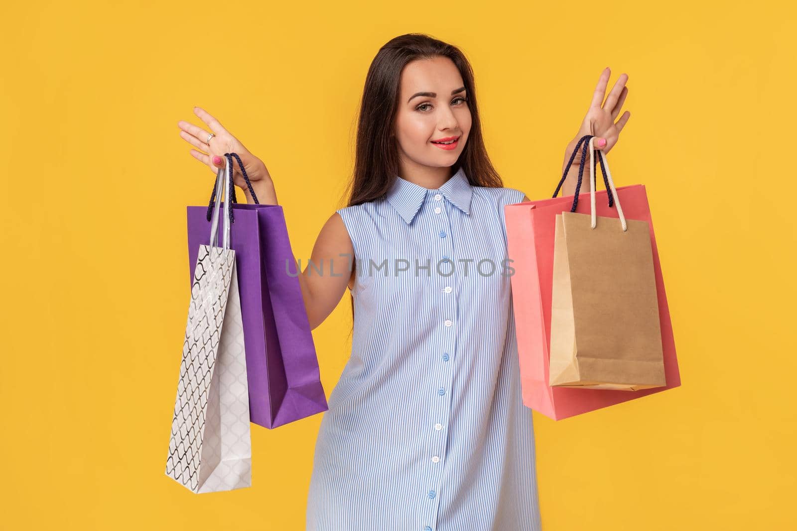 Image of a young brunette lady in white and blue summer striped dress posing with shopping bags and looking at camera over yellow background. Shopping. Sale