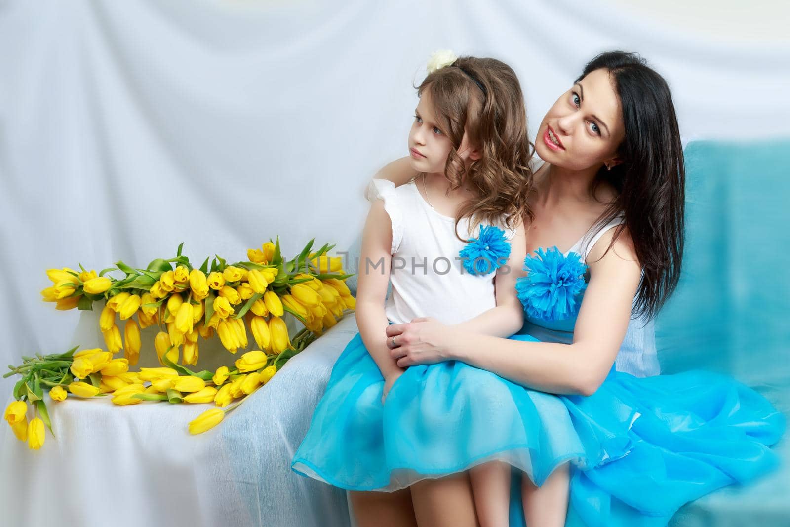 Beautiful young mother and little daughter in the same blue dresses on the sofa with a bouquet of flowers.