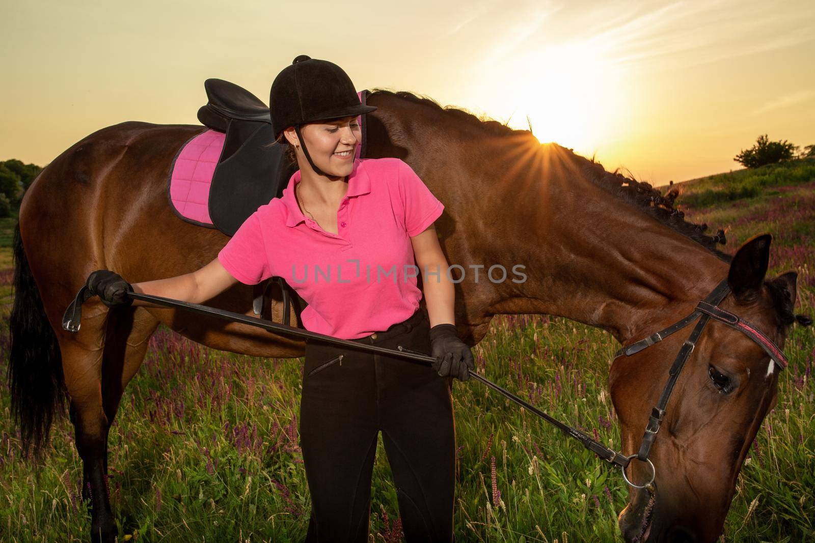 Beautiful smiling girl jockey stand next to her brown horse wearing special uniform on a sky and green field background on a sunset. Equitation sport competition and activity.