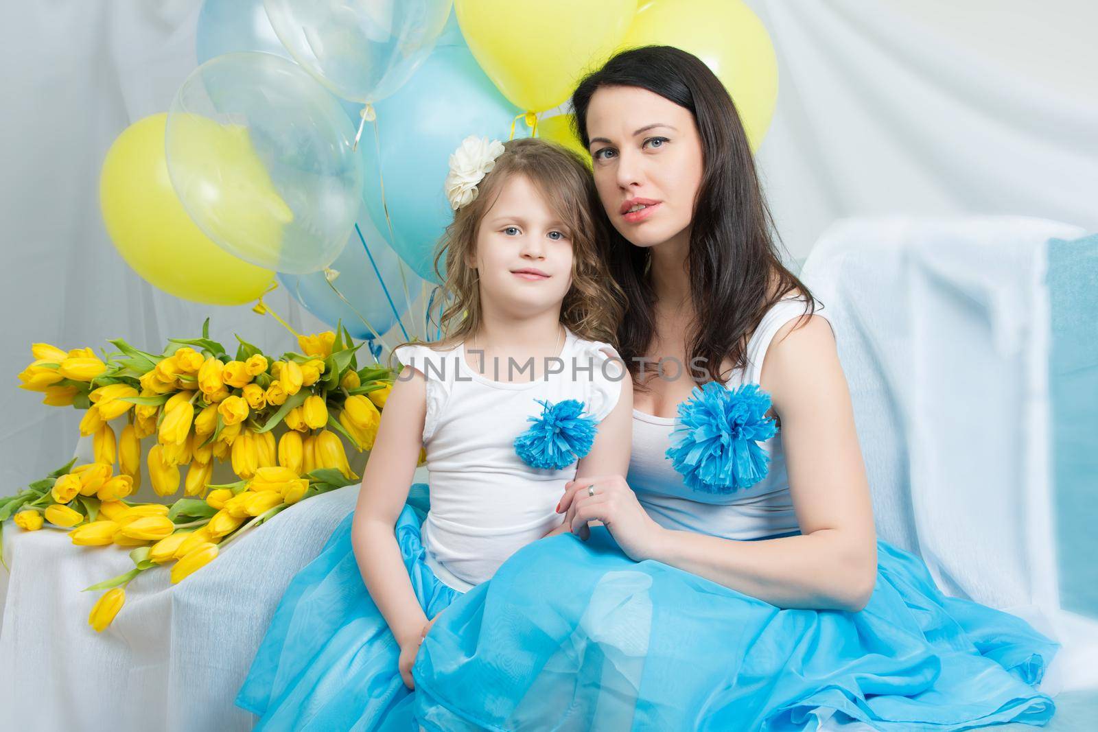 Beautiful young mother and little daughter in the same blue dresses on the sofa with a bouquet of flowers.