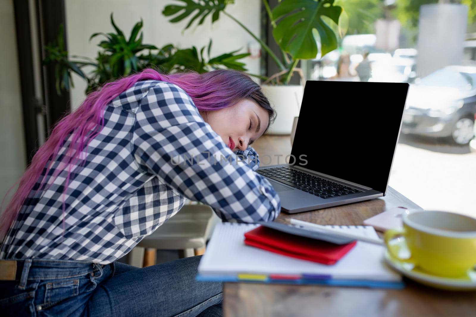 Young woman lying on her arms on the table in cafe in front of laptop with cup of coffee, sleepy. Rear shot of female student learning online via laptop before her lectures.