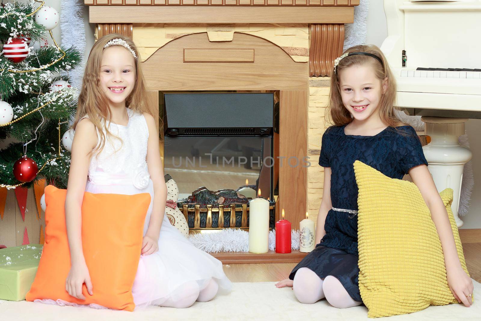 Cute little twin girls, sitting on the floor near the Christmas tree and electric fireplace on which candles are burning. In the hands of the girls boxes with gifts.