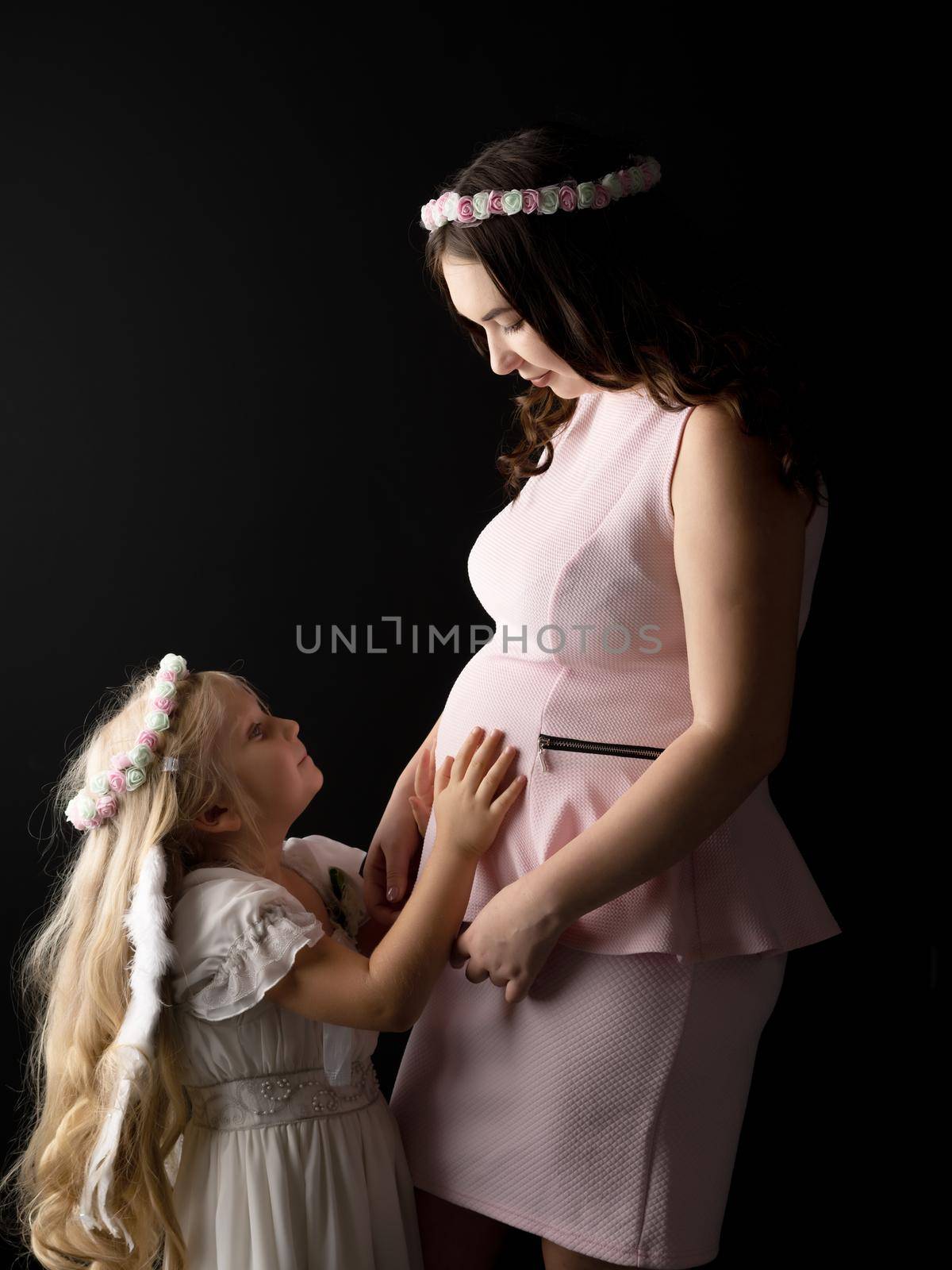 A little girl in the image of an angel hugs the belly of her eldest pregnant sister. The concept of family happiness, the birth of a child.