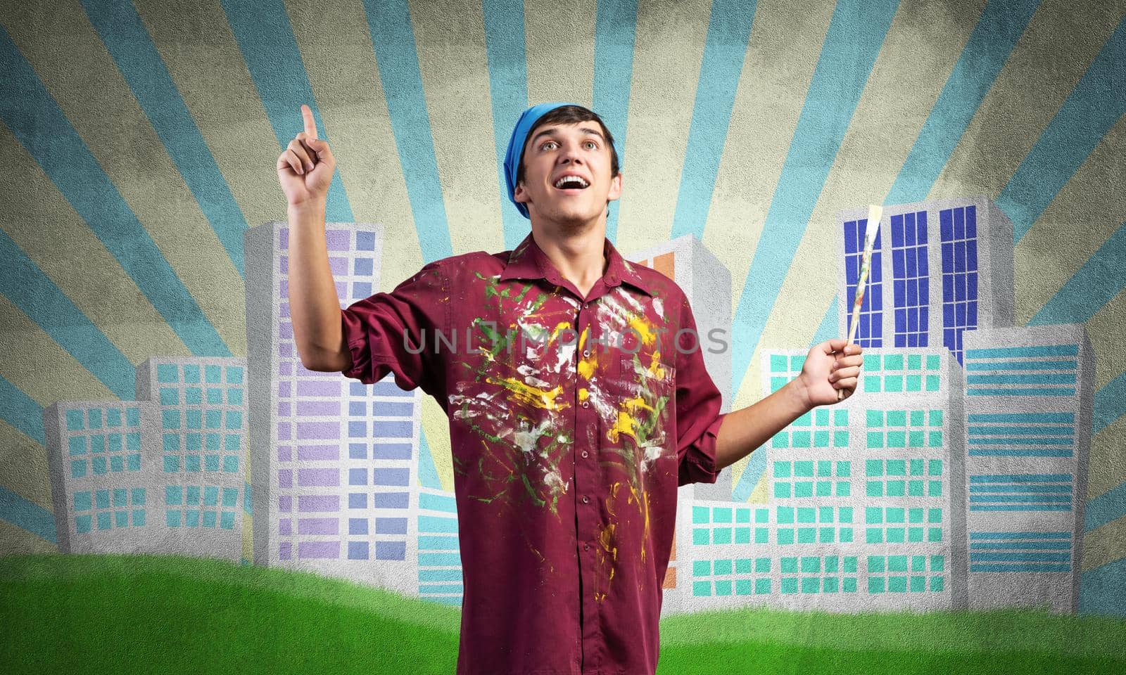 Young male artist holding paintbrush. Happy painter in shirt and bandana standing on background colorful picture on wall. Modern cityscape with high skyscrapers artwork. Creative hobby and profession