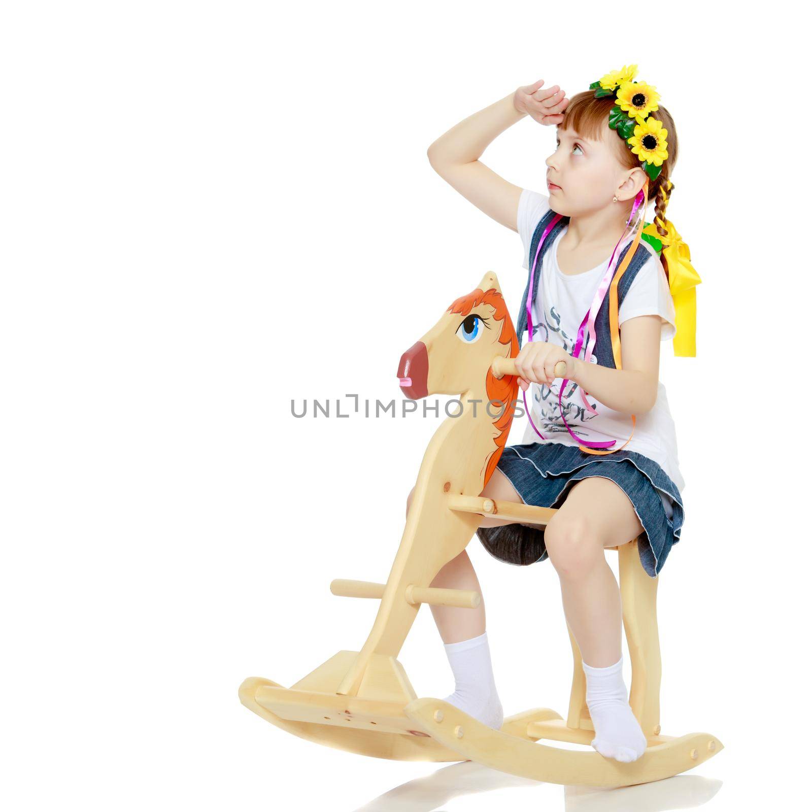 A beautiful little blonde girl in a summer short dress, and a wreath of yellow flowers on her head.The girl is rocking on a wooden horse.
