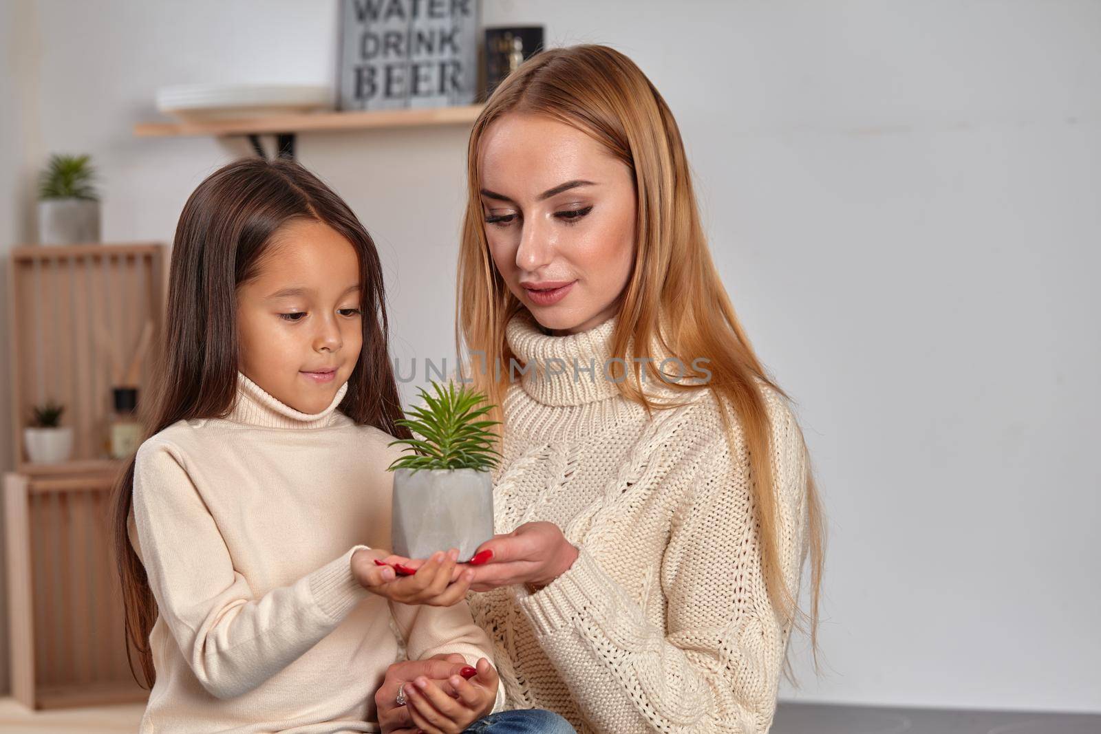 Beautiful adorable good-looking cheerful mom and her small little daughter on weekend, sitting on countertop, consider a flower in a pot in light kitchen