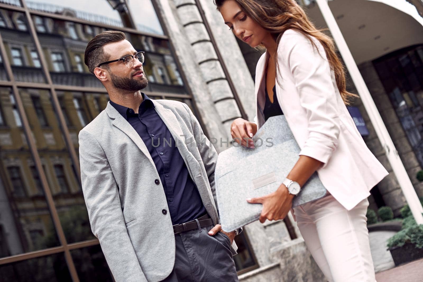 Waiting for colleague. Two young business people standing outside on the city street man looking curious at woman taking documents from her bag close-up by friendsstock