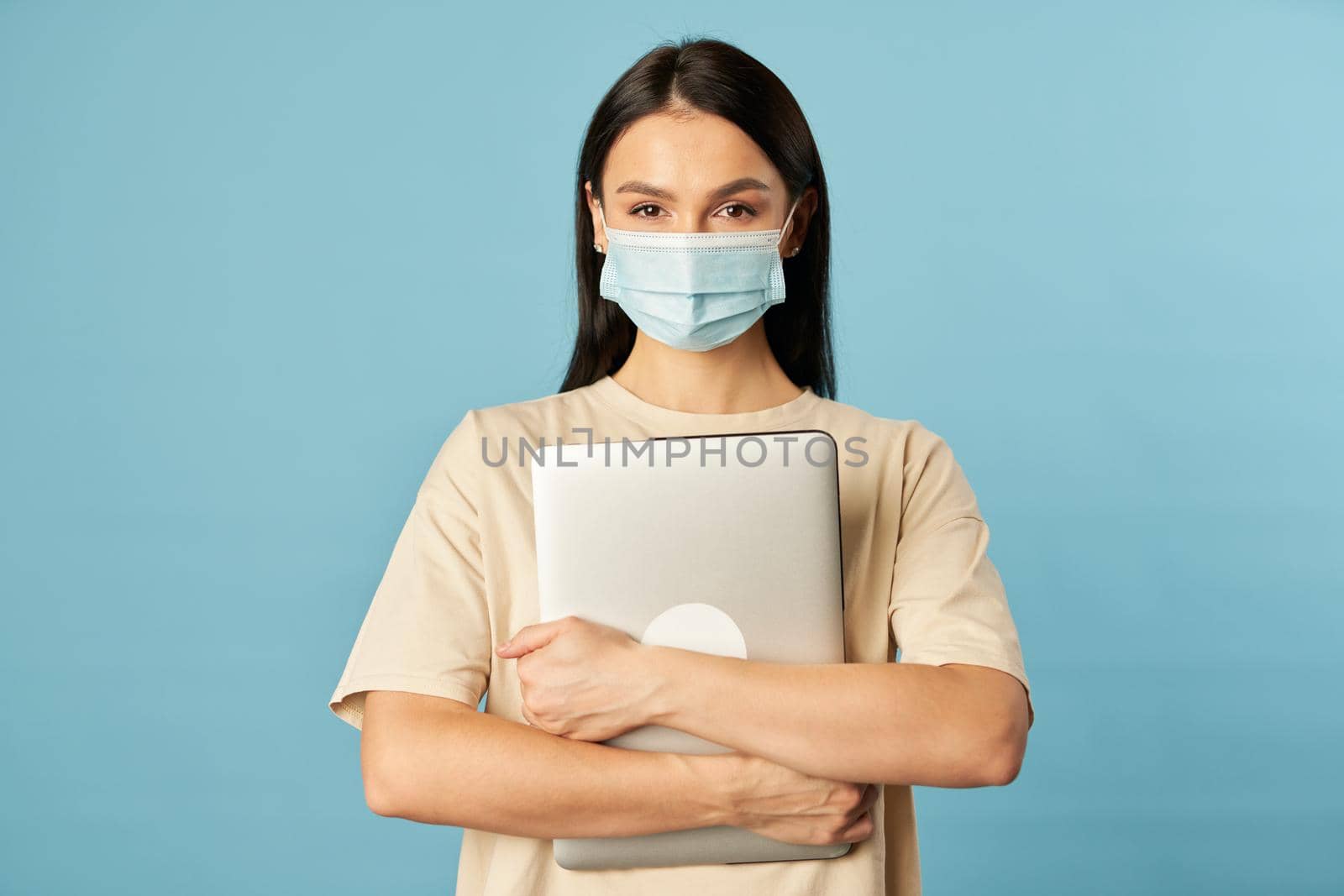 Waist up of young woman wearing face mask and holding laptop while looking at camera, isolated on blue background. Copy space. Quarantine, epidemic concept