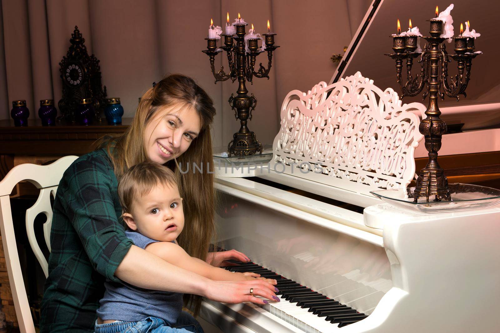 A young mother with a small child on Christmas night with candles near a large white piano. The concept of holidays and family happiness.