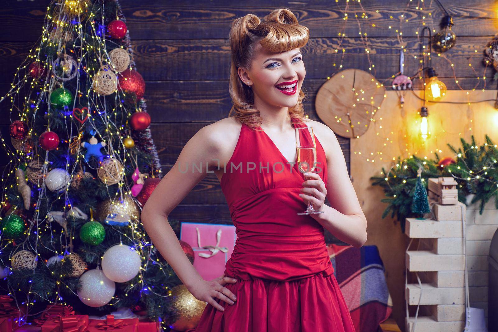 Smiling ponup woman in evening red dress with glass of sparkling wine over home Christmas tree background. Fashion and beauty make-up and hairstyle for Christmas