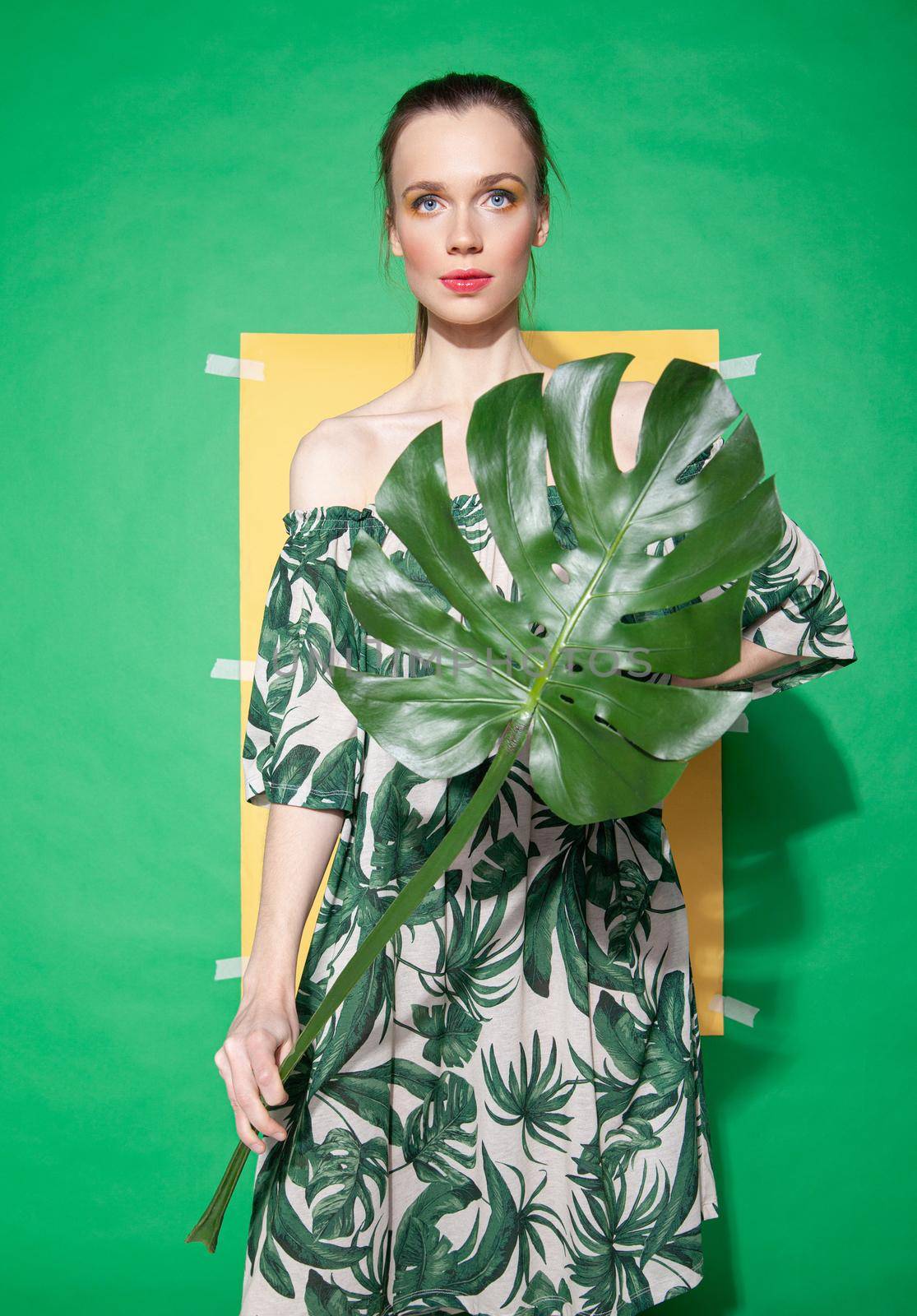 Young female model in stylish dress with floral ornament holding leaf while standing against yellow paper sheet on green background in summer