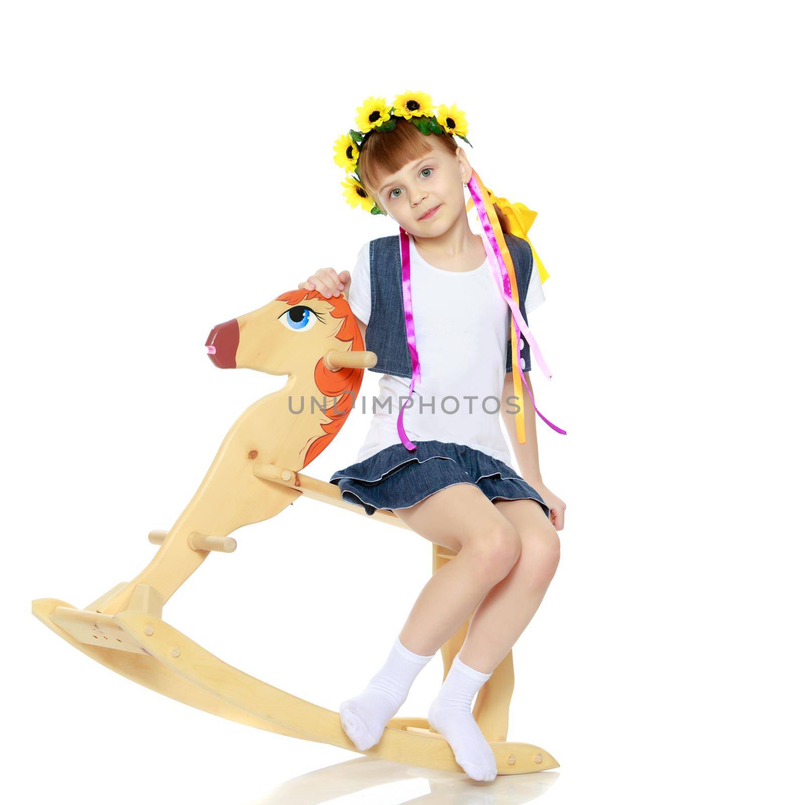 A beautiful little blonde girl in a summer short dress, and a wreath of yellow flowers on her head.The girl is rocking on a wooden horse.