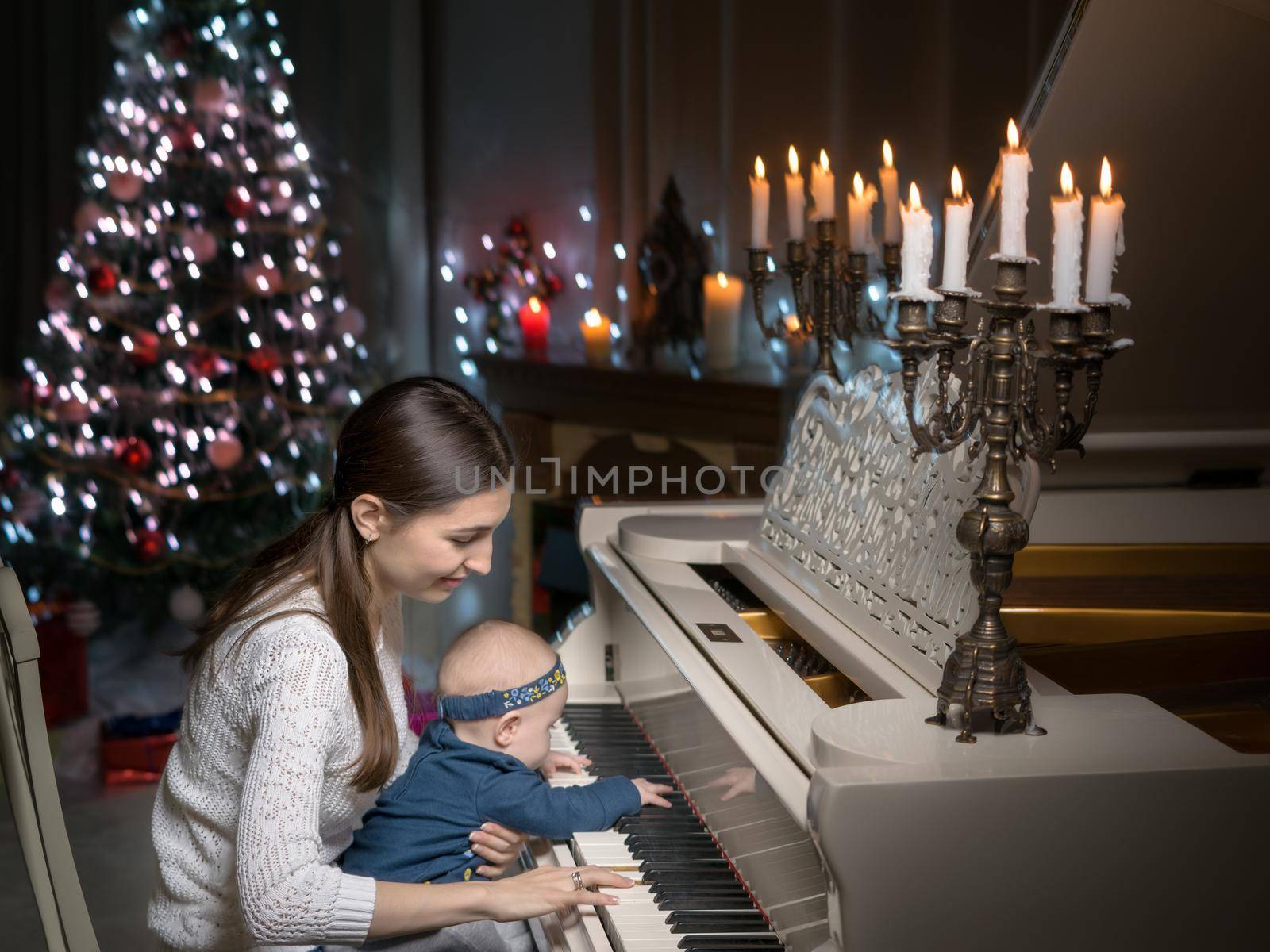 A young mother with a small child on Christmas night with candles near a large white piano. The concept of holidays and family happiness.