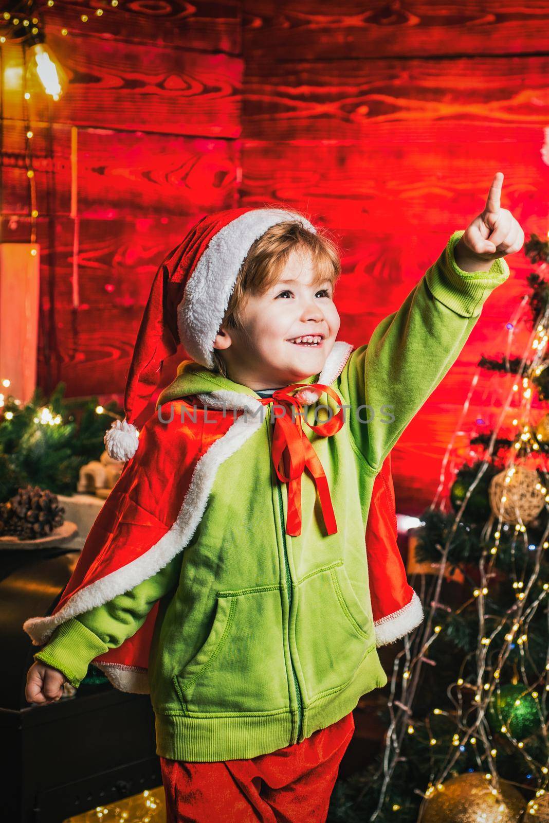 Magic Christmas night for kids. Happy excited smiling boy with pointing finger by the Christmas tree. Little kid is wearing Santa clothes