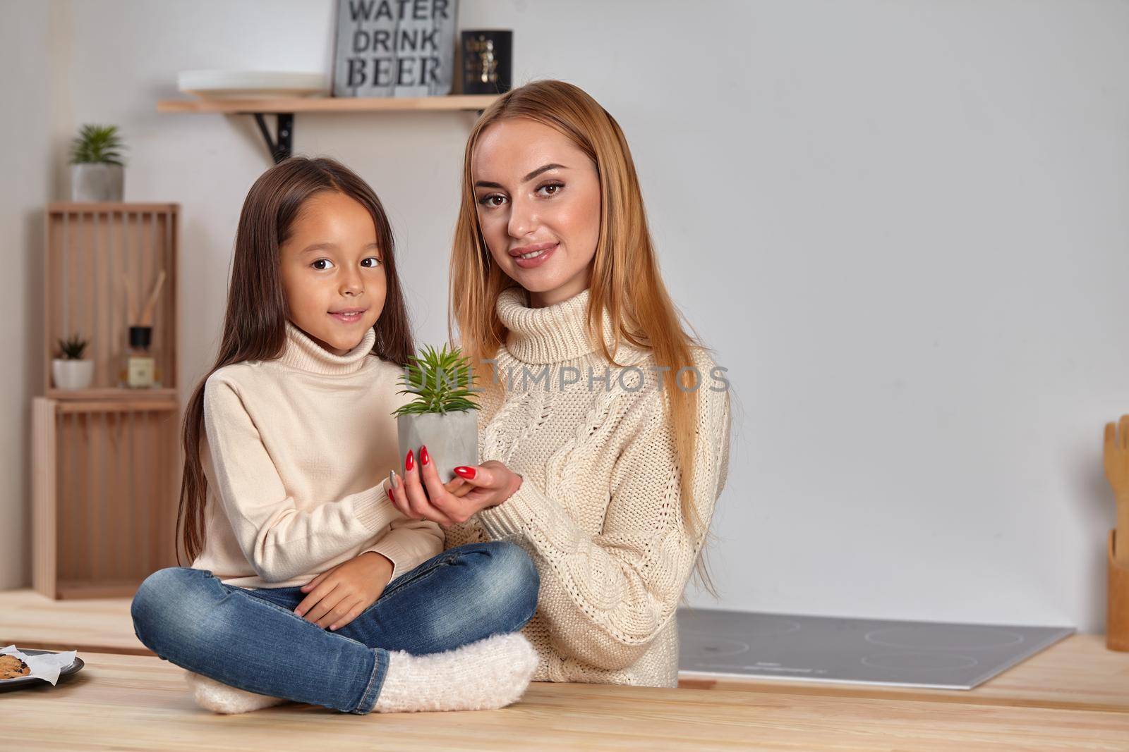 Beautiful adorable good-looking cheerful mom and her small little daughter on weekend, sitting on countertop, consider a flower in a pot in light kitchen