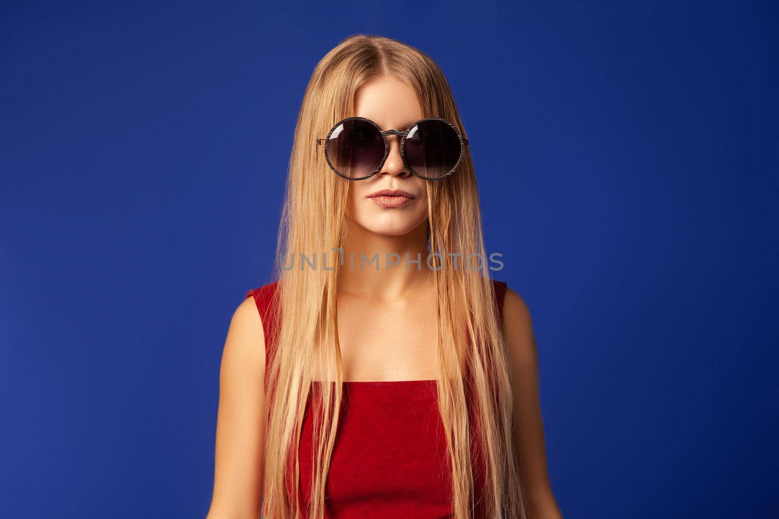 Young female model with long blond hair wearing stylish sunglasses against blue background