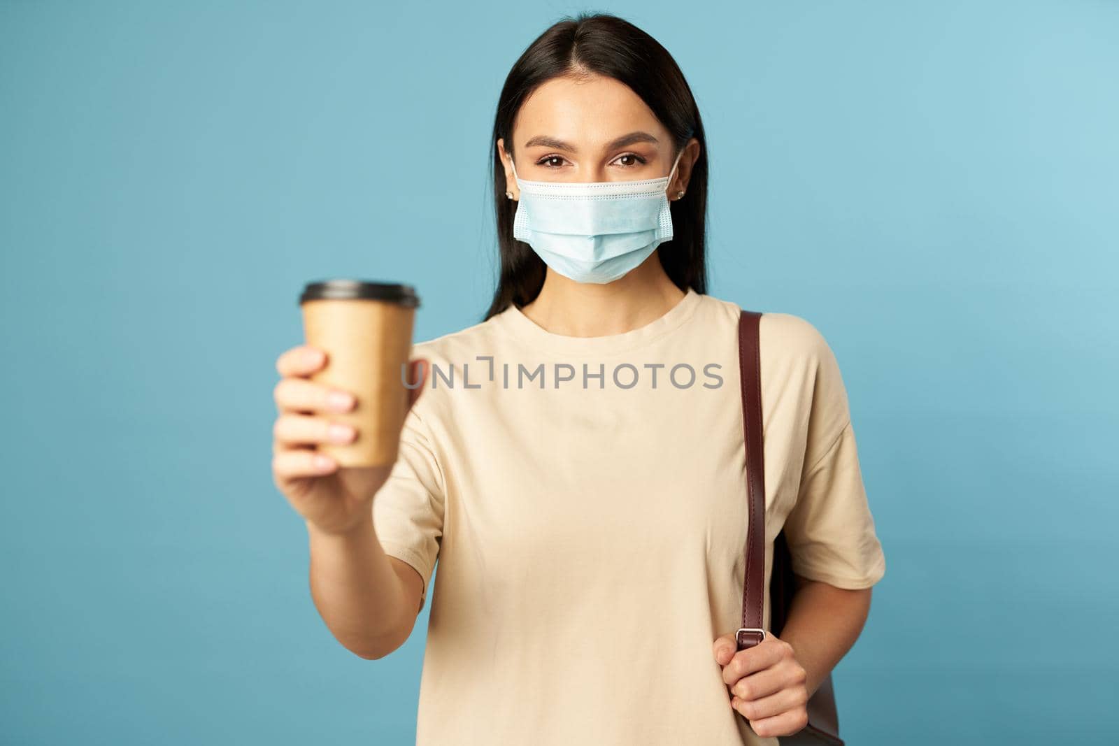 Happy young female wearing face mask posing with hot drink and backpack while posing in studio against blue background. Copy space. Quarantine, epidemic concept