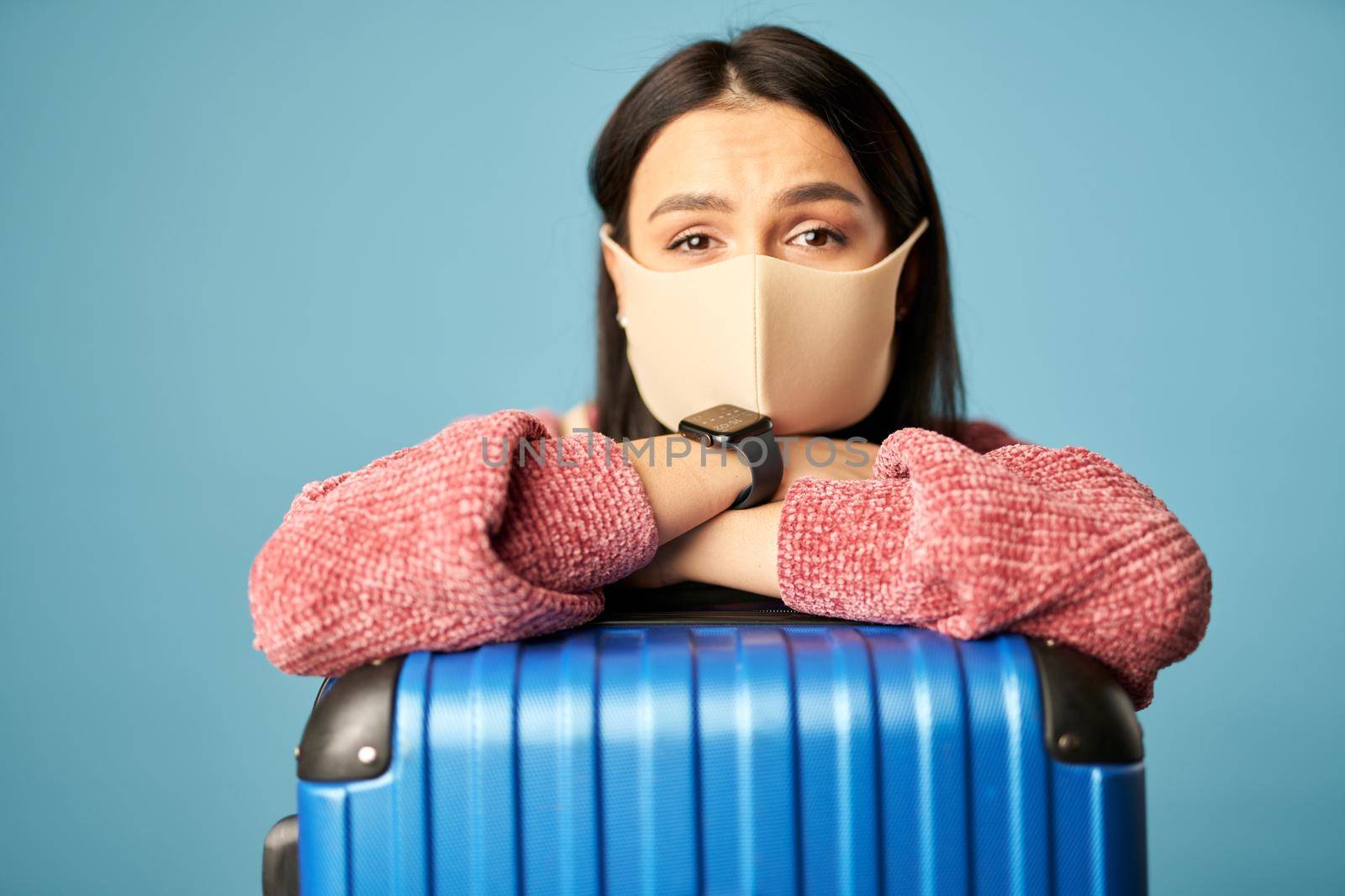 Pretty lady wearing protective mask with suitcase posing against blue background. Copy space. Concept of travel, coronavirus